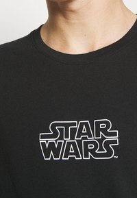 Musta puuvillapaita, jossa on valkoinen "STAR WARS" -logo. Pyöreä kaula-aukko, lyhyet hihat, sileä rakenne ja rento istuvuus.