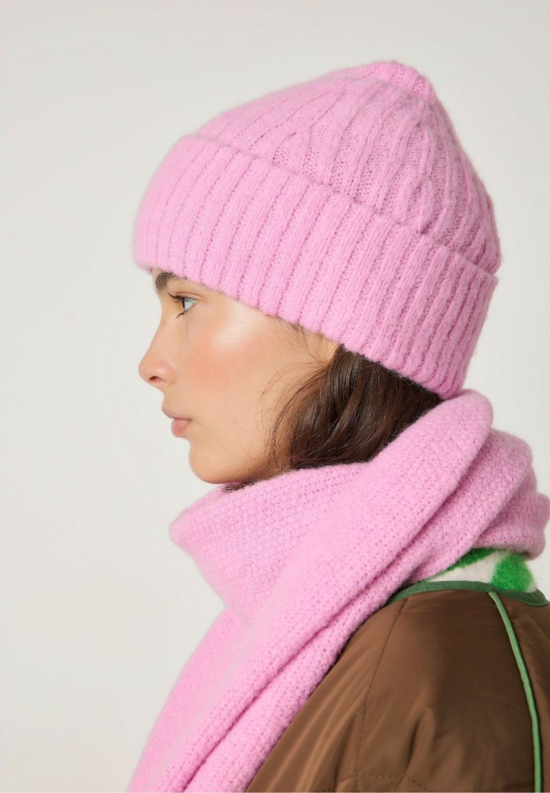 Roosa kudumine beanie ribakujulise mustriga ja pehme tekstuuriga, kantud koos samanäolise roosa salliga, koos pruuni väliskattega.