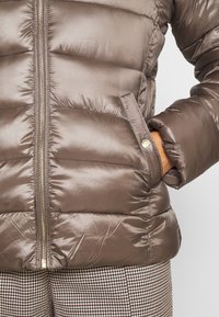 Hnědá péřová bunda s lesklou prošívanou texturou, předním zipem a bočními kapsami. Obsahuje kulatou kovovou logo ozdobu na kapse.