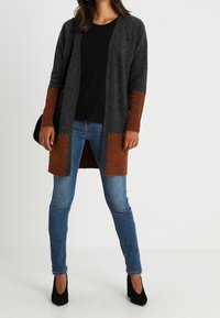 Stickad kardiagan i mörkgrått med rostfärgade ärmar. Öppen framsidan, långa ärmar, kombinerad med blå jeans och svarta klackar.