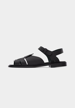 Black leather open-toe sandal with adjustable ankle strap and flat sole, showcasing textured material and side cutout details.