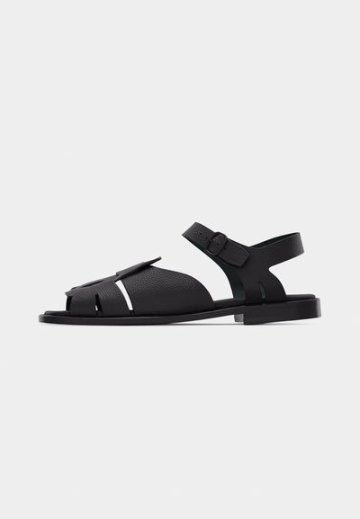 Sandale ouverte en cuir noir avec bride de cheville réglable et semelle plate, présentant un matériau texturé et des détails de découpe latérale.