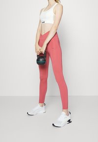 Reggiseno sportivo bianco leggero con logo, leggings rosa e kettlebell nero. Le scarpe da ginnastica sono bianche con accenti neri e sovrapposizioni testurizzate.