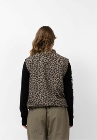 Een bruin en zwart leopardenprint vest met een cropped ontwerp en zonder mouwen, gecombineerd met een zwart shirt met lange mouwen eronder.