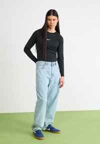 Person with long black hair wears black Nike long-sleeve top, light blue loose jeans, and blue Nike sneakers standing on green floor.