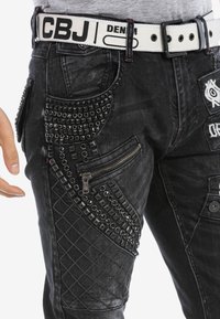 Zwarte denim broek met spikes en studs, een ritssluitingzak, een gewatteerde textuur en een gevlochten riem met de branding "CBJ DENIM".
