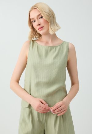 Kobieta z krótkimi blond włosami, ubrana w bezrękawnik w jasnym odcieniu zieleni oraz pasujące spodnie, stojąca z delikatnie złożonymi rękami.