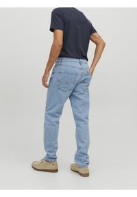 Muž v světlých modrých džínách, tmavě modrém tričku a béžových semišových botách, stojící s jednou rukou v zadní kapse na jednoduchém pozadí.