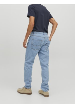 Hombre vestido con jeans de color azul claro, camiseta azul marino y zapatos de ante beige, de pie con una mano en el bolsillo trasero contra un fondo liso.