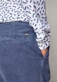Personne portant un pantalon en velours côtelé bleu avec une étiquette sur la poche arrière "BOSS" et une chemise blanche avec des motifs abstraits bleus, main dans la poche latérale.