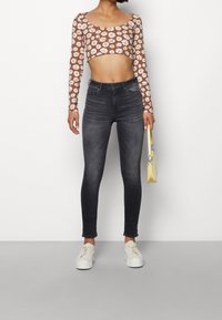 Mujer con jeans skinny de cintura alta en negro desgastado, zapatillas blancas, una blusa corta de manga larga marrón con margaritas blancas, sosteniendo un pequeño bolso amarillo.