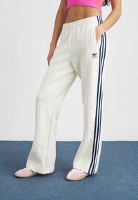 Mulher vestindo calças brancas de ponto de cable da Adidas com riscas azul marinho e um suéter cropped rosa, de pé sobre um tapete azul.