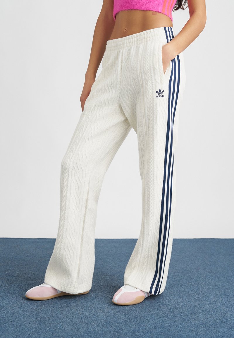 Mulher vestindo calças brancas de ponto de cable da Adidas com riscas azul marinho e um suéter cropped rosa, de pé sobre um tapete azul.