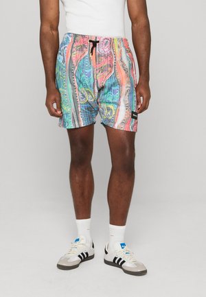 Man draagt meerkleurige korte broek met patroon en zwarte trekkoorden, witte sokken en witte sneakers met zwarte strepen, staand tegen een effen achtergrond.