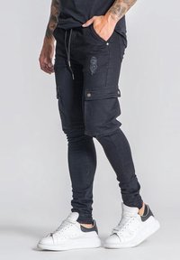 Joggers cargo negros con un ajuste slim, presentan un pequeño detalle de rasgado, bolsillos en los laterales y una cintura con cordón. Combinados con zapatillas blancas.