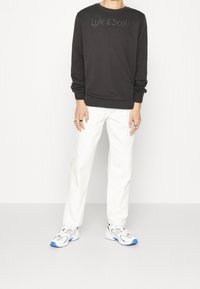 Svart sweatshirt med präglad logotyp och rund halsringning, matchad med vita raka byxor och vita sneakers med blå detaljer.