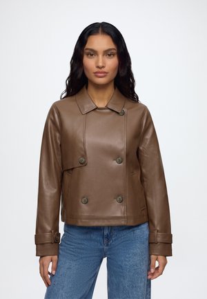 Femme aux longs cheveux foncés portant une veste en cuir marron à double boutonnage et un jean bleu sur un fond blanc uni.
