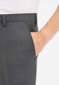 Hand inserted into the front pocket of tailored dark gray pants worn with a white shirt against a plain white background.