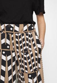 Wide-leg trousers in a patterned fabric featuring black and white geometric designs against a tan background. Side pockets visible.