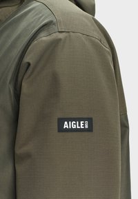 Manche de veste verte avec un patch rectangulaire noir cousu portant l'inscription "AIGLE 1853" sur le haut du bras, sur un fond uni.