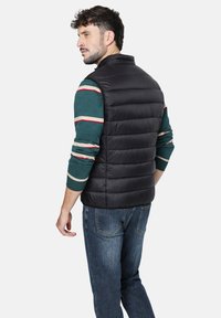 Chaleco acolchado negro con cuello alto, que presenta secciones acolchadas y un diseño ligero. Lleva puesto un shirt de manga larga a rayas multicolores.