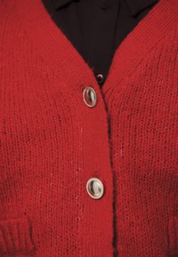 Lähedalt vaadatuna punane kootud kardigan, millel on kaks metallist ovaalset nööpi musta rõiva peal.