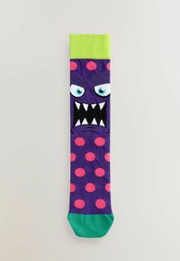 Calcetín morado con lunares rosas, puño verde y una cara de monstruo con dientes negros afilados y ojos azules. Hecho de material elástico.