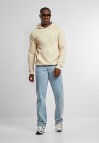 Hoodie beige chiaro con tasca a marsupio, abbinato a jeans azzurri e sneakers bianche con dettagli grigi, modello che indossa occhiali da sole.