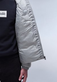 Uomo che indossa pantaloni neri e un felpa blu navy, mentre tiene aperta una giacca a vento argento-grigia che mostra la fodera interna liscia e il bordo della cerniera.