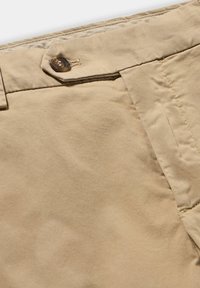 Pantaloni di cotone beige con chiusura a bottone, frontale piatto e texture liscia, con cuciture visibili e un aspetto leggermente stropicciato.