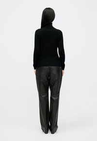 Mulher com cabelo longo e escuro veste um suéter preto de gola alta e calças de couro pretas largas, de pé voltada para longe contra um fundo simples.