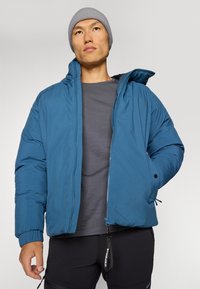 Blå puffjacka med ståkrage, frontdragkedja och elastiska manschetter. Bärs över en grå t-shirt, accessoriserad med en grå stickad mössa.