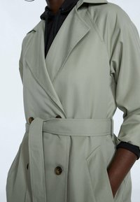 Trench coat verde claro con diseño de doble botonadura, cinturilla y bolsillos laterales. Cuenta con un cuello con muesca y una textura suave.