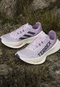 Ζεύγος ανοιχτόμωβ παπούτσια trail running Adidas Terrex τοποθετημένα σε πετρώδες έδαφος με χορτάρι και πέτρες γύρω.