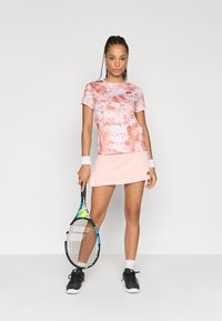 Saia de ténis rosa claro combinada com uma t-shirt multicolorida com um padrão em espiral. Ténis desportivos pretos. Segurando uma raquete de ténis com uma bola.