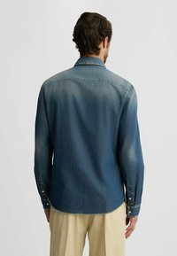 Camisa de mezclilla azul oscuro con un degradado desvanecido, puños con botones y un dobladillo curvado. Presenta un collar clásico y una textura suave.