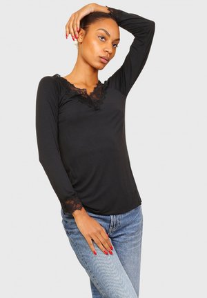 Haut noir à manches longues avec un col en dentelle et des poignets, fabriqué à partir d'un tissu lisse. Le mannequin pose sur un fond gris. Les jeans sont de couleur bleu clair.
