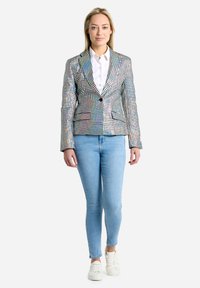 Živahna blazer jakna v pixelastem vzorcu z enim gumbom, zarezanim ovratnikom in dvema sprednjima žepoma, v kombinaciji s svetlo modrimi oprijetimi jeans hlačami.