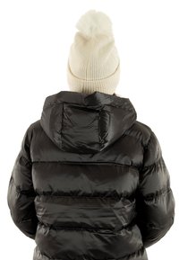 Cappotto piumino nero con cuciture orizzontali e cappuccio, abbinato a un berretto in maglia color crema con un pompon sulla cima.