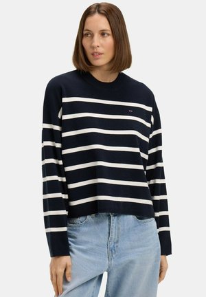 Femme aux cheveux bruns mi-longs, portant un pull bleu marine à rayures horizontales blanches et un jean bleu clair, sur fond blanc.