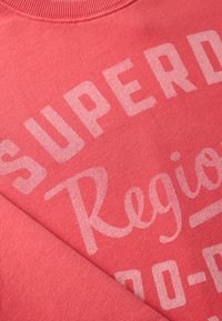 Felpa corallo con colletto a costine e grande testo bianco che recita "SUPERD" e "Regione", realizzata in un tessuto morbido e testurizzato.