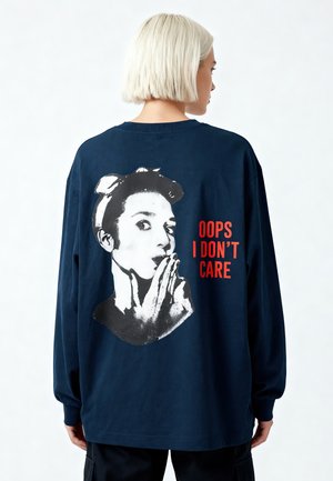 Persona bionda vista da dietro che indossa una felpa blu navy con una grafica di volto in bianco e nero e testo rosso con la scritta "OOPS I DON'T CARE."