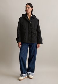 Zwarte waterdichte jas met een capuchon, vooraan met drukknoopsluiting, twee grote zakken en een zachte kraag. Gepaard met wijde denim broeken en schoenen.