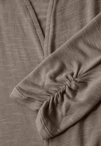 Brun, strukturerad tygblus med V-ringning och rynkade ärmar. Materialet verkar mjukt med ett subtilt ribbat mönster.