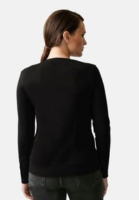 Schwarzes Langarmshirt mit rundem Ausschnitt, glatter Textur und tailliertem Design. Kombiniert mit dunklen Jeans, die dekorative Gesäßtaschen aufweisen.