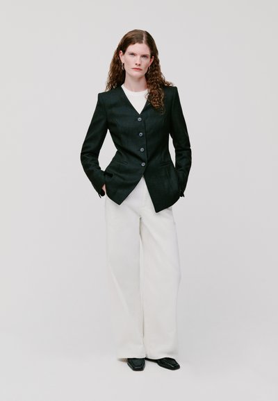 Blazer nero su misura con vita aderente, design monopetto e grandi bottoni, abbinato a pantaloni bianchi larghi. Tessuto liscio.
