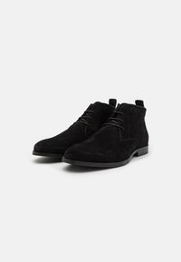 Botine negre din nubuc, cu vârful rotund, prezentând un design cu șireturi și un toc jos, plasate pe un fundal alb.