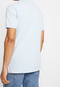 Ljusblå kortärmad polo med en mjuk textur, ribbad krage och rak fåll, bärs tillsammans med denimjeans. Bakvy visas.