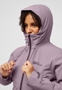 Giacca impermeabile viola con colletto alto, cappuccio regolabile e logo del marchio sul petto. Presenta una chiusura con zip e dettagli in presa.