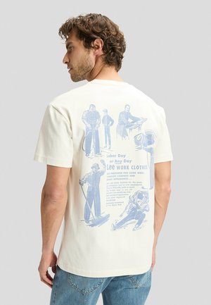 Mann in hellem T-Shirt mit Vintage-blauen Illustrationen und Text über Lee Arbeitskleidung, von hinten gezeigt, trägt hellblaue Jeans.